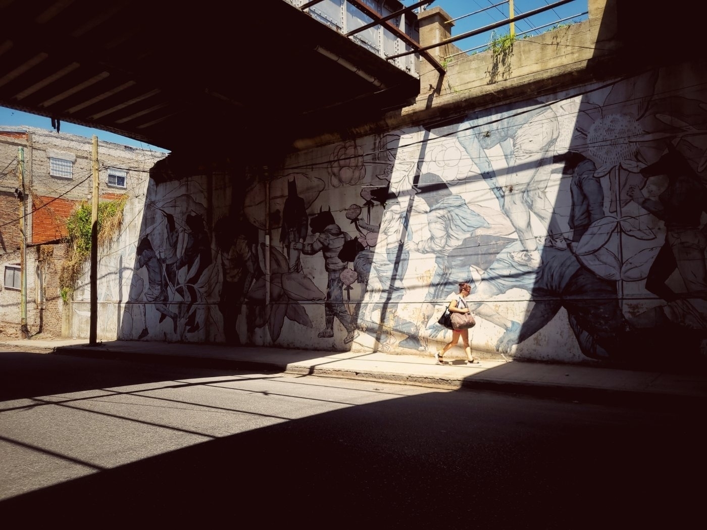 A person walks along a sunlit pathway beneath a bridge adorned with mural art.