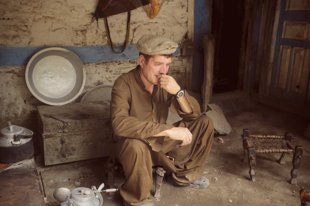 A person wearing traditional attire and a hat is sitting pensively in a rustic room with kitchenware and a woven stool.