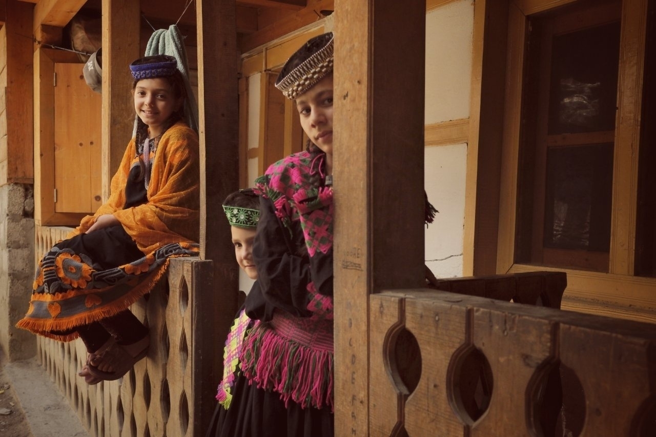 Three children dressed in colorful traditional attire are sitting and standing on a wooden porch.