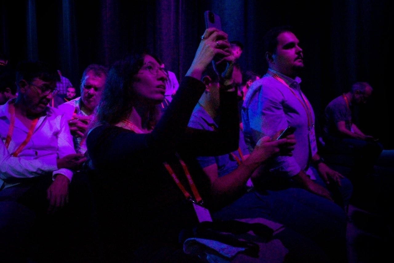 People seated in an audience capture photos or interact with their phones in a dimly lit setting.