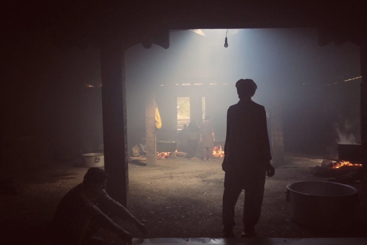 In a dimly lit room with shafts of light filtering through, a person stands silhouetted in front of a fire, while another sits nearby.