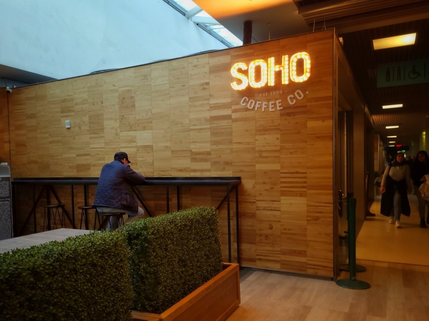 A person is sitting alone at a counter in a coffee shop called "SOHO Coffee Co." with a wooden interior and some greenery nearby.