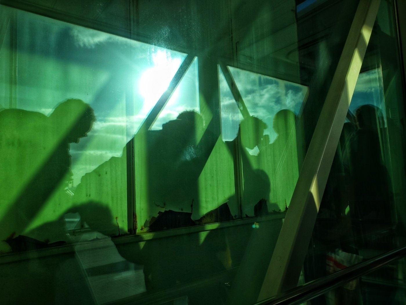 Silhouetted figures are seen through green-tinted glass with sunlight streaming in and clouds visible in the background.