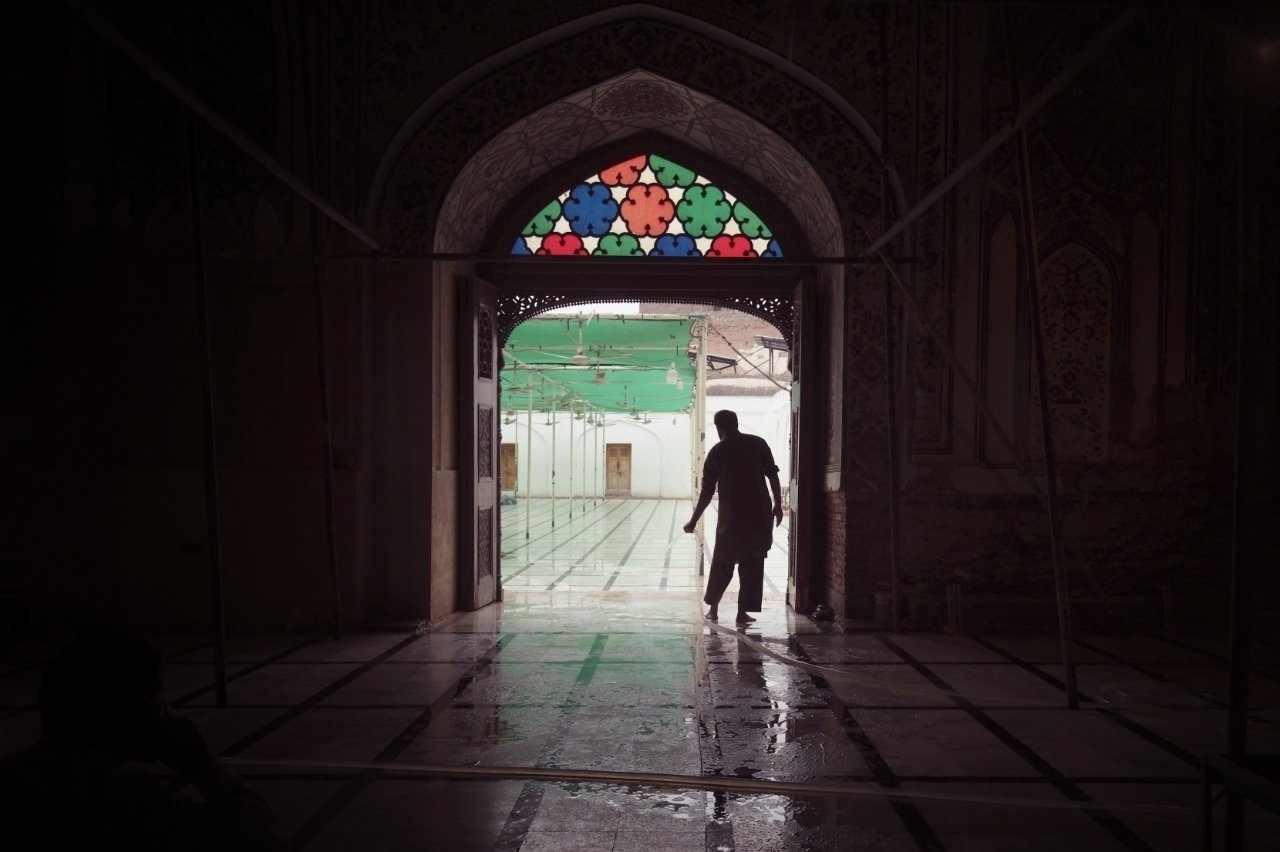 A person walks through the arched doorway of a dimly lit building, leading to a tiled outdoor area with a colorful stained glass design above the entrance.