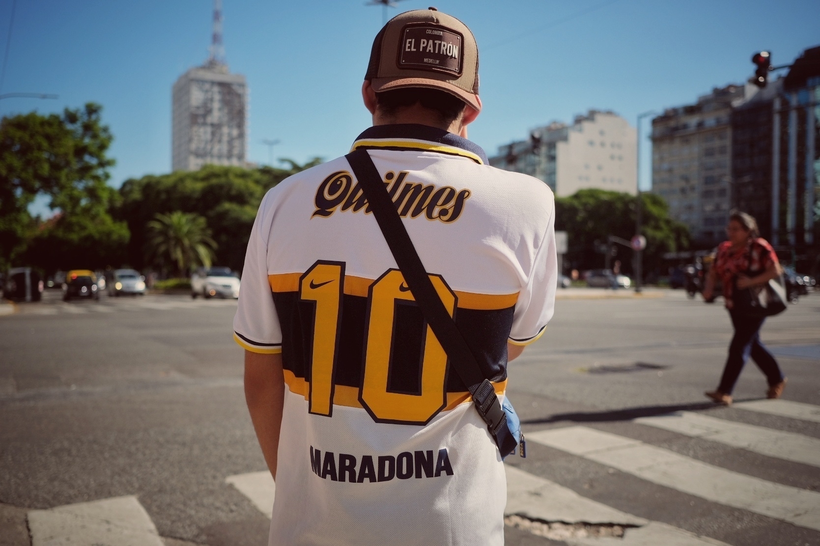 A person wearing a "Maradona 10" sports jersey and a cap stands at a crosswalk in an urban setting.