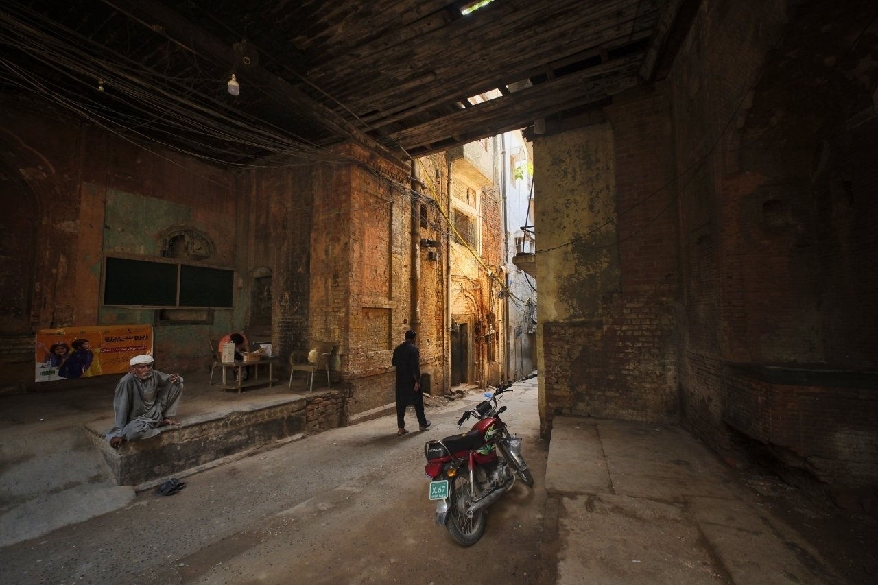 A dimly lit, rustic alley features an old motorcycle, a few people seated and standing, and weathered brick architecture.