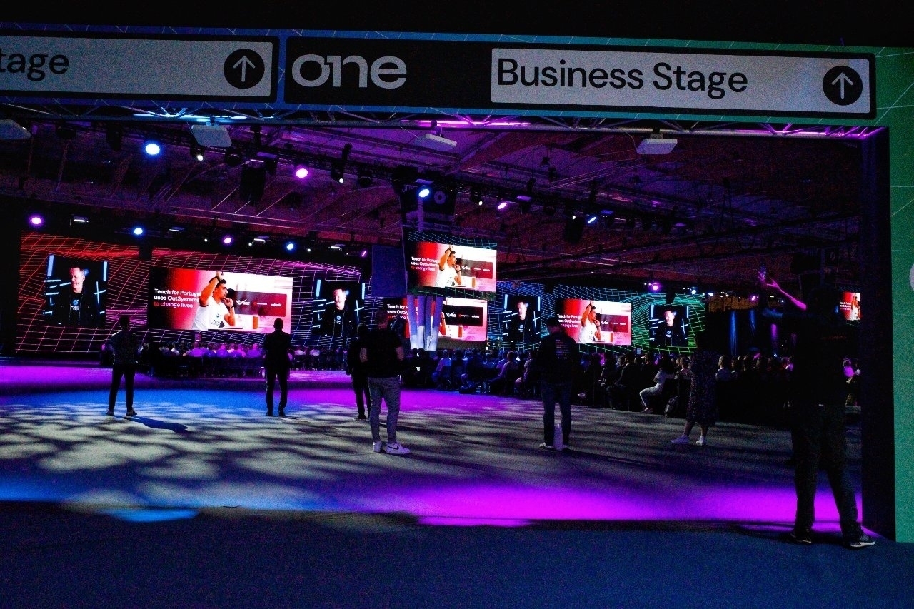 People are gathered in a venue with colored lights and large video displays under a banner labeled Business Stage.