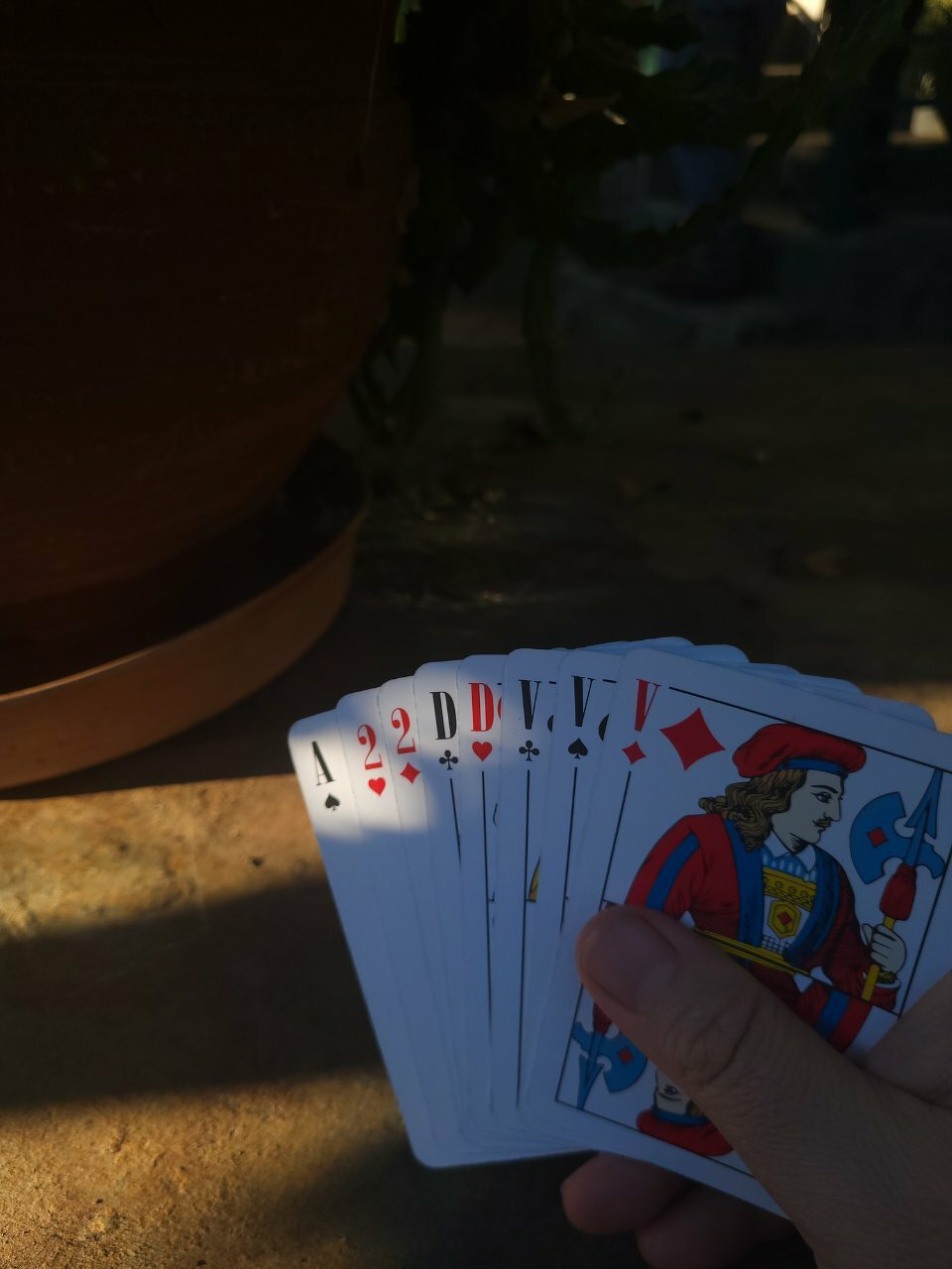 A hand is holding a fan of playing cards, including the ace and number two of hearts, alongside a king of diamonds, set against a shaded outdoor backdrop.