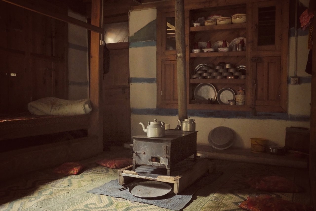 A rustic kitchen features a central wood-burning stove with teapots on top, surrounded by traditional kitchenware and a cozy seating area with cushions on the floor.