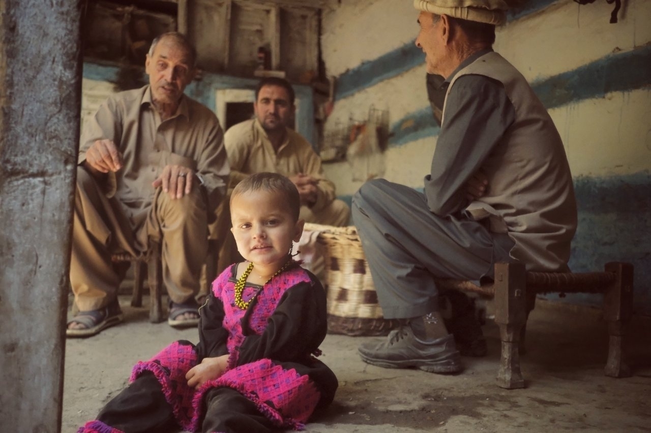 A small child in a traditional outfit sits on the floor, while three adults are gathered in conversation inside a rustic room.