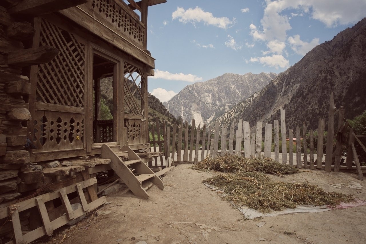 A rustic wooden house with a lattice design sits next to a wooden fence, set against a backdrop of majestic mountains.