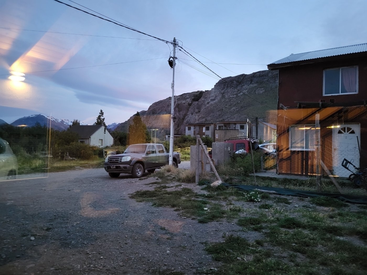 A rural scene features a pickup truck parked on a dirt road, with houses and rocky hills in the background, under a dusky sky.