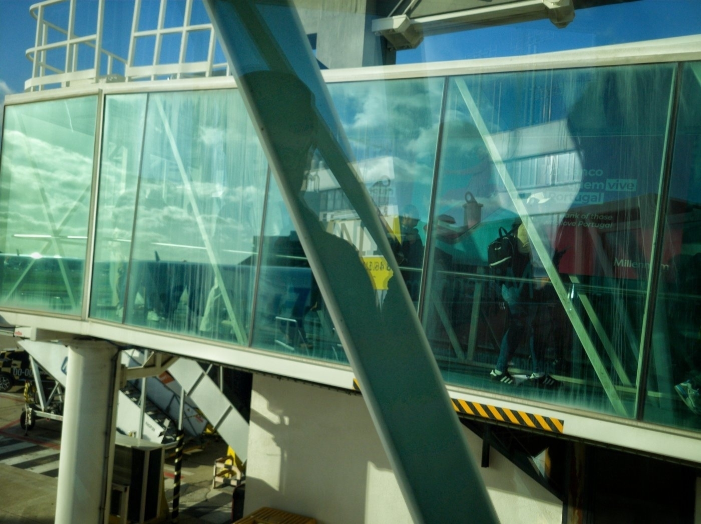 A glass-walled airport jet bridge is seen with reflections of people and blue skies.