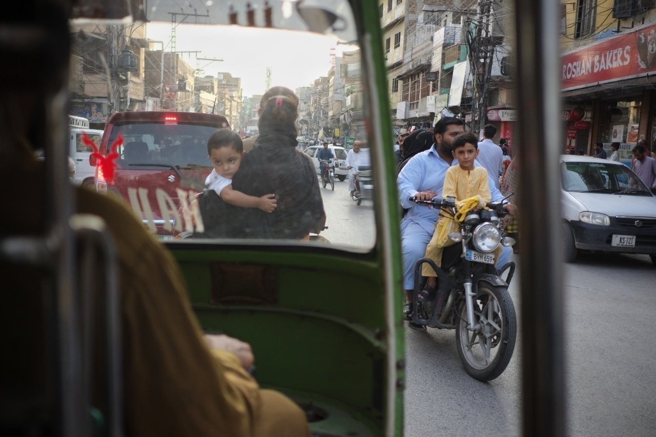 A busy street scene shows a woman and child in a tuk-tuk while a man and child ride a motorbike alongside mixed traffic.