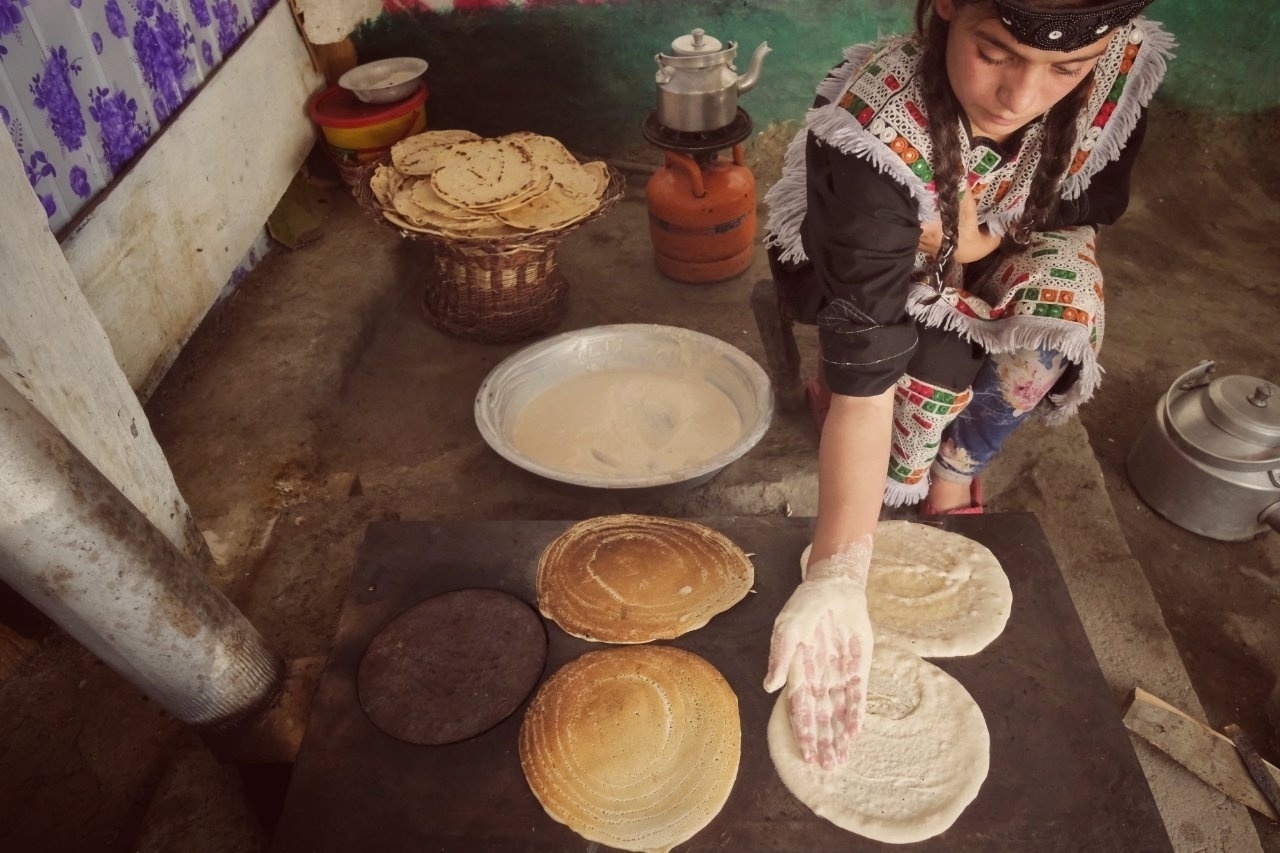 A woman is preparing traditional flatbreads on a cooking surface with various kitchen items around her.