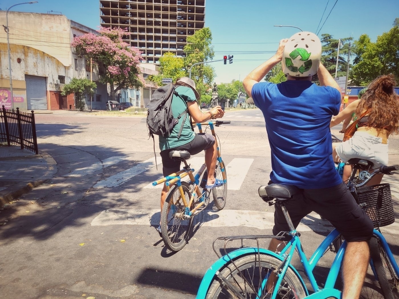 People on bicycles wait at a street crossing in an urban setting with trees and buildings around.