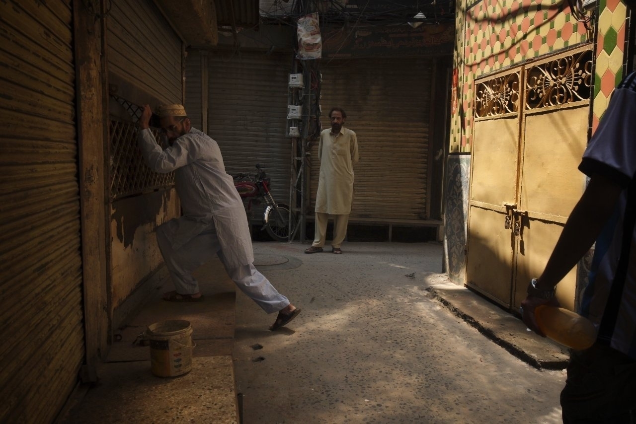 A person in traditional attire is pushing a metal shutter in a dimly lit alley, with another person standing in the background and a third person at the edge holding a bottle.