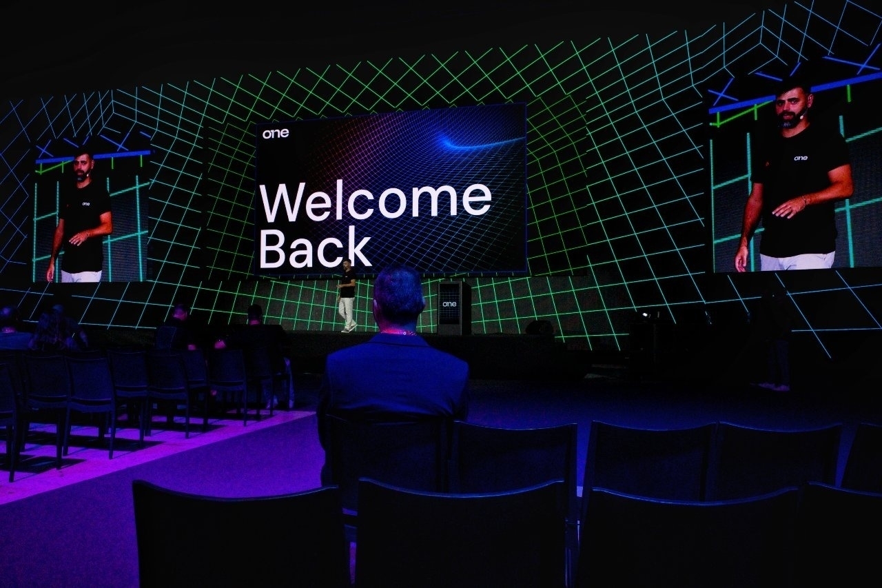 A man is standing on a stage with a large screen displaying the words Welcome Back in a venue with a grid-patterned backdrop.