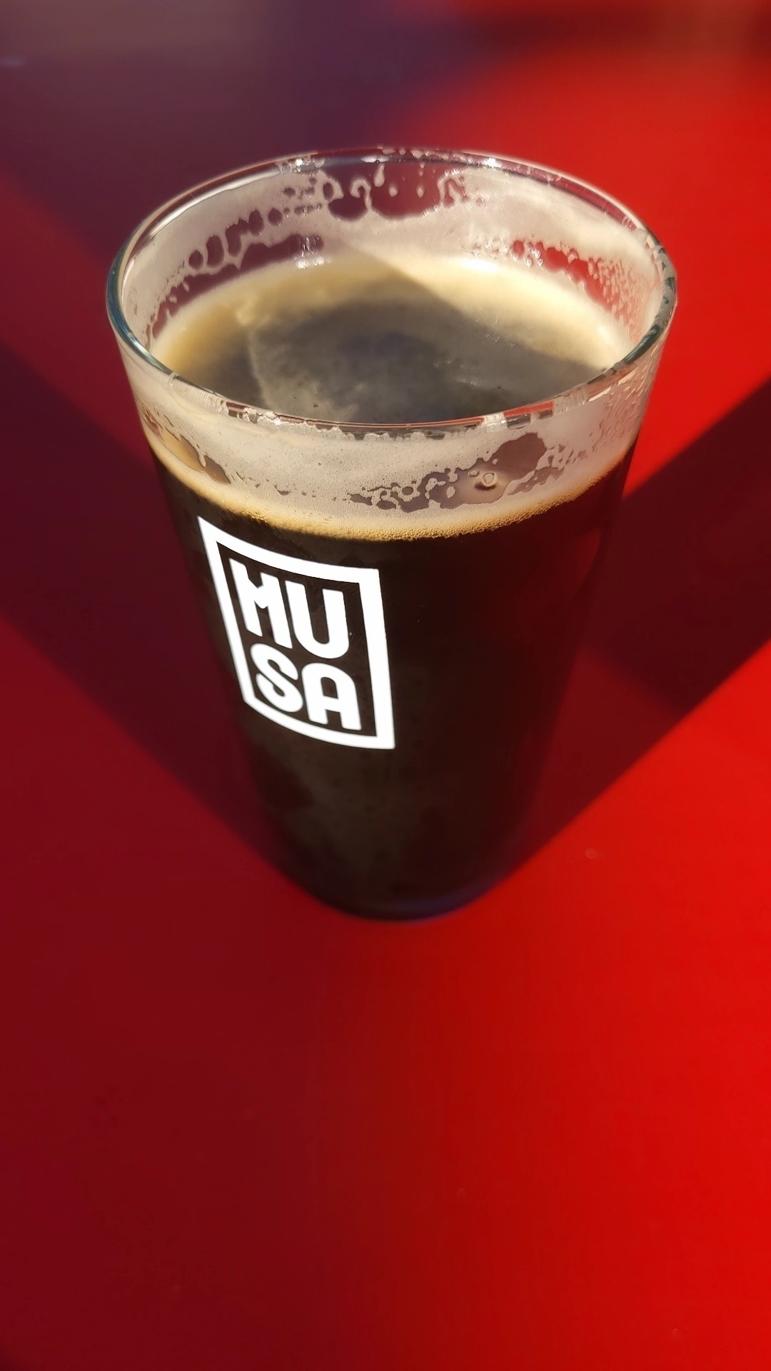 A glass of dark beer with a frothy head sits on a bright red surface, featuring a "HUSA" logo.