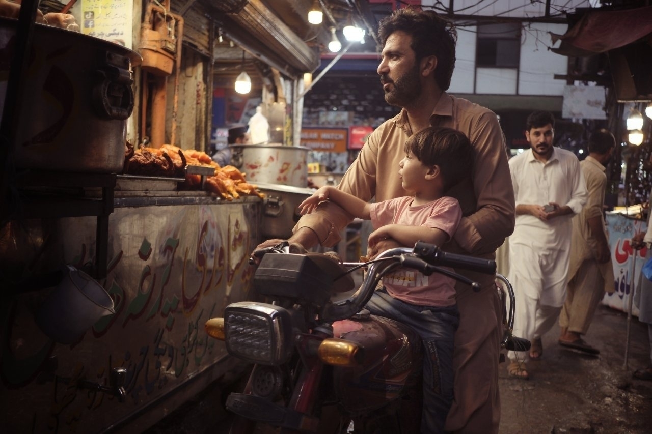 A man and a child on a motorcycle stop to look at food stalls in a bustling market.