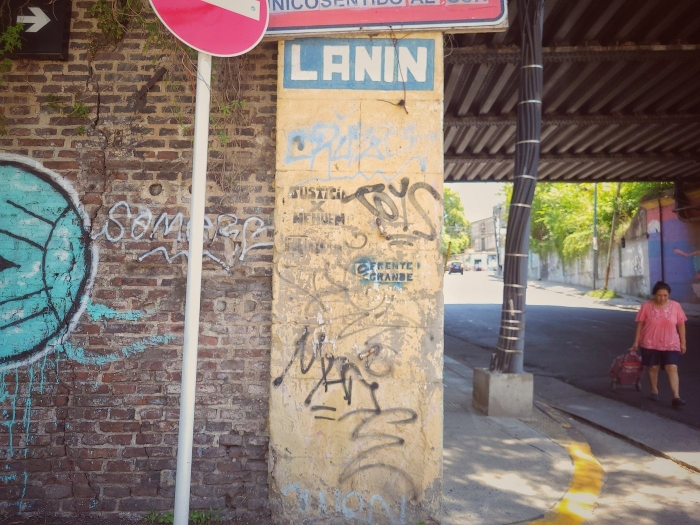 A graffiti-covered street scene features a brick wall, a signpost, and a person walking on the sidewalk under a bridge.