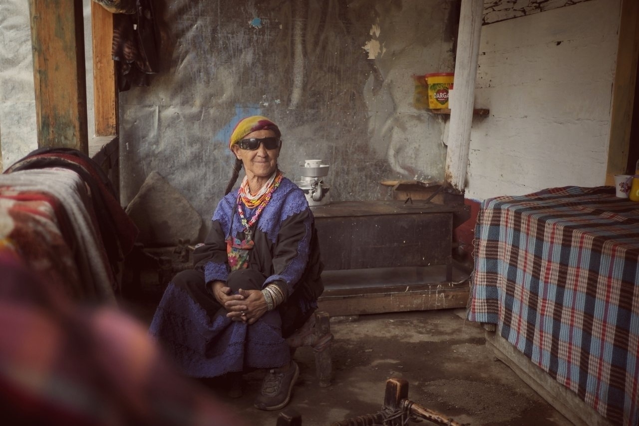 An older person wearing colorful clothing and sunglasses sits contentedly inside a rustic, cozy space with patterned textiles.