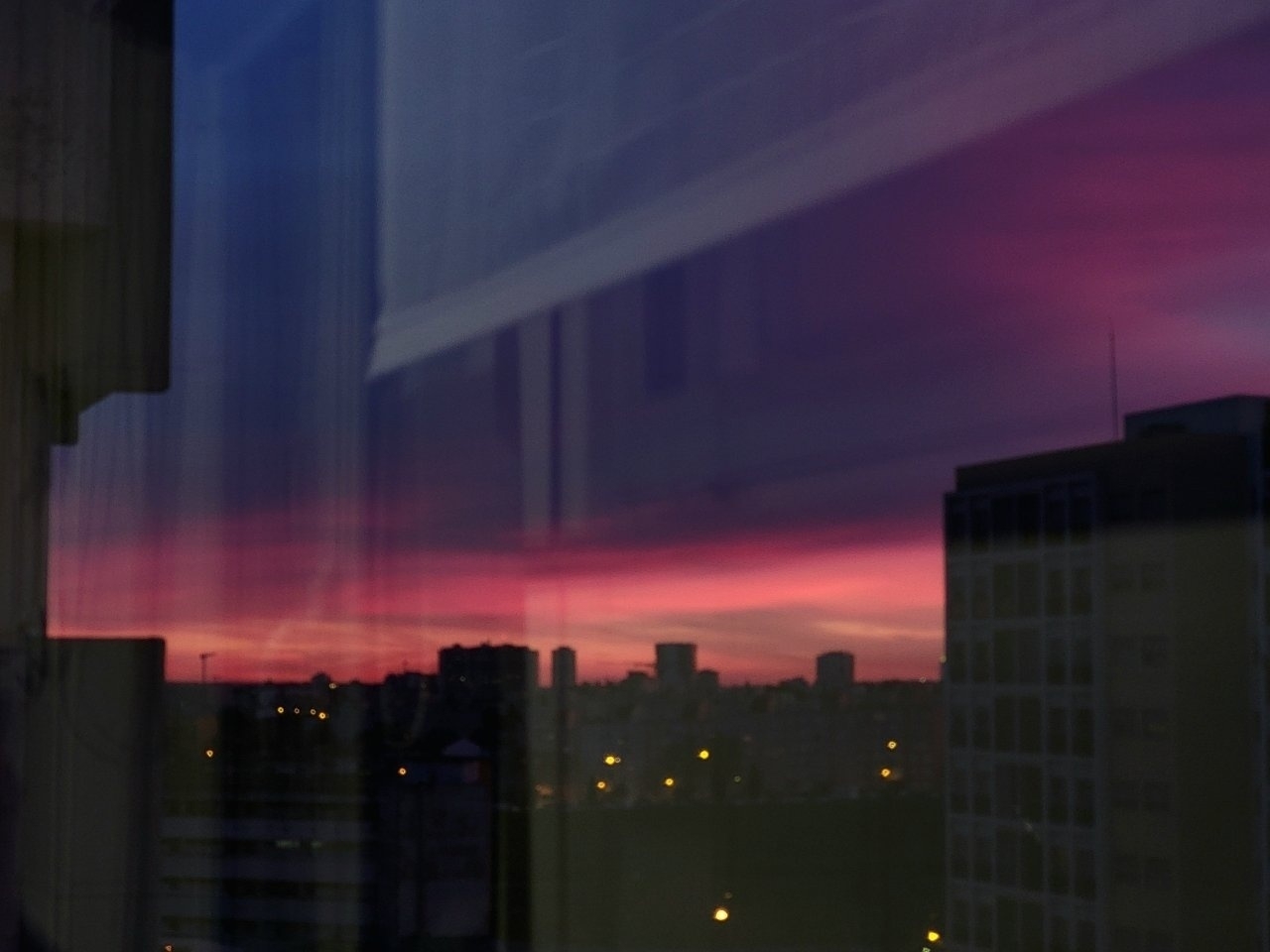 A cityscape at dusk is reflected in a window, showcasing a vibrant purple and pink sky above silhouetted buildings.