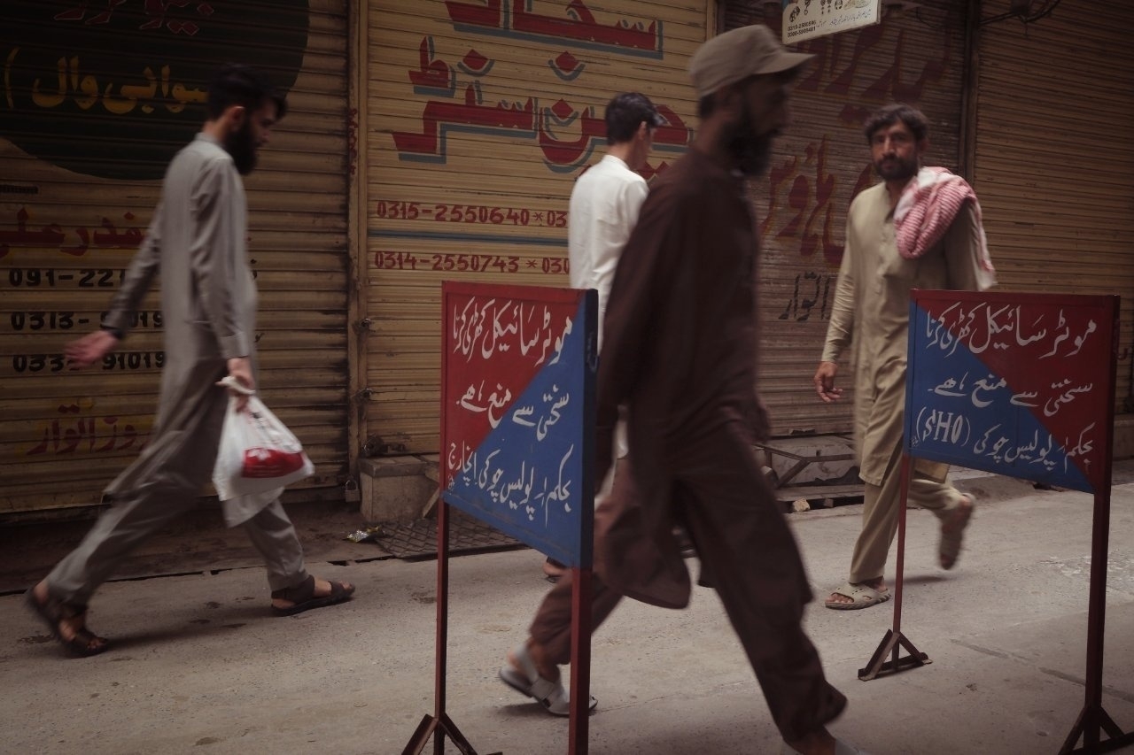 Men walk down a narrow street with metal barriers displaying text in a non-English script, in front of closed shops.