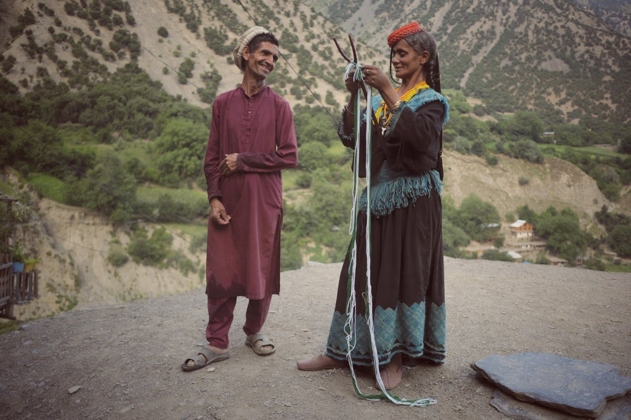 Two people in traditional clothing stand on a rocky terrain with mountains and greenery in the background, engaged in an activity with strings or ropes.