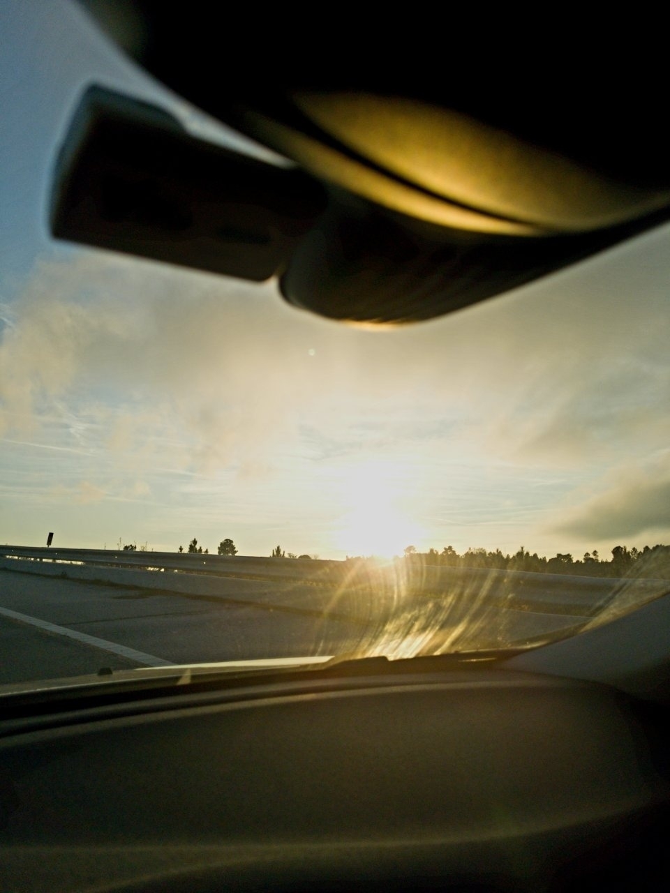 A bright sunset view is seen from inside a car, with road and sky visible through the windshield.