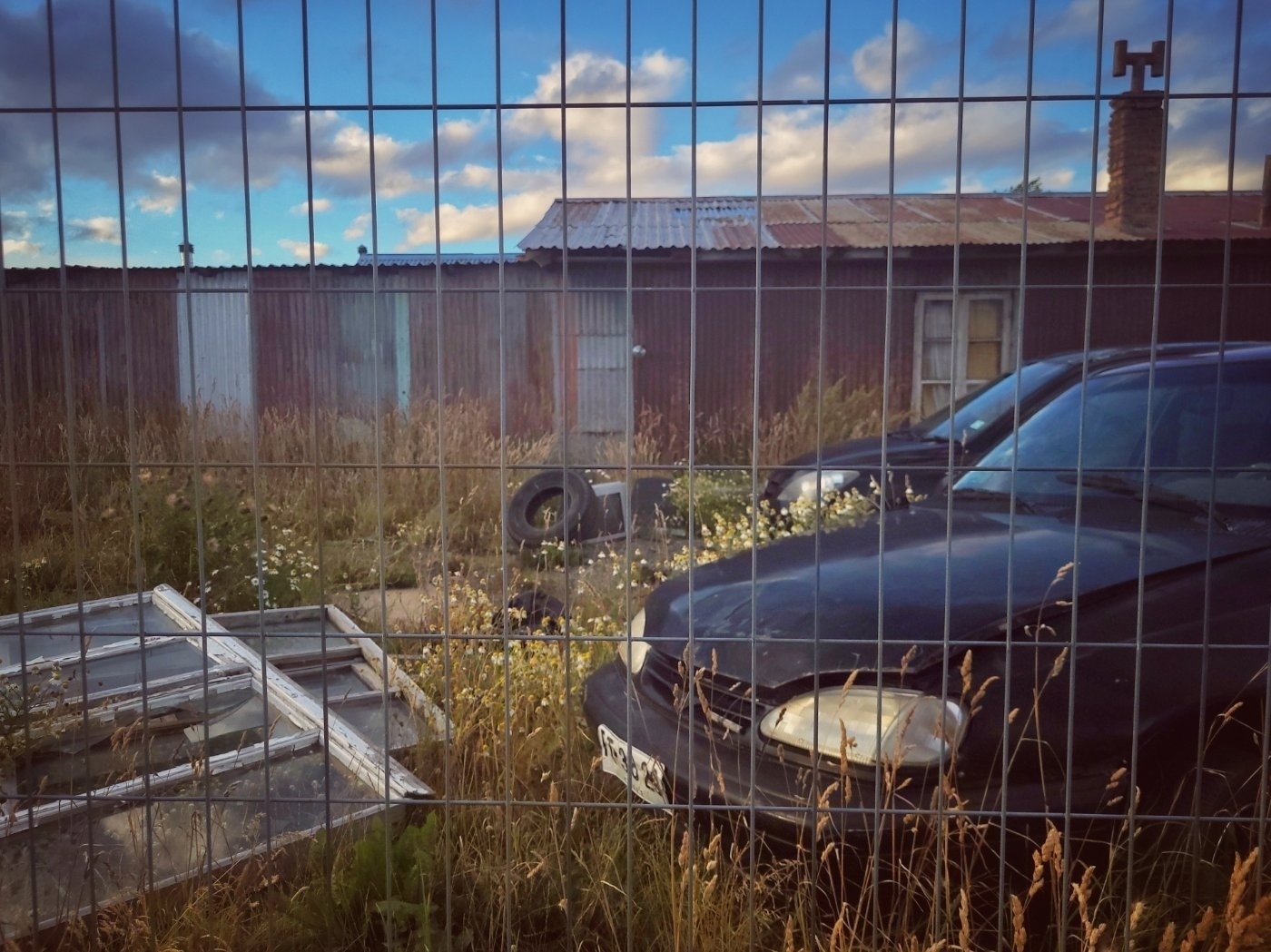 A fenced area contains overgrown grass, an old building, scattered debris, and parked cars.