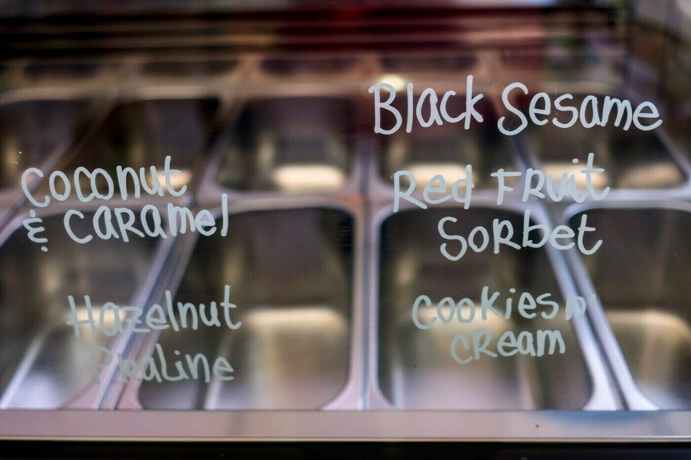 Ice cream flavors such as Coconut & Caramel, Hazelnut Praline, Black Sesame, Red Fruit Sorbet, and Cookies & Cream are written on a glass display in front of empty metal containers.