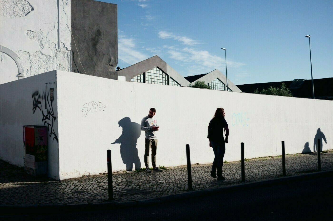 Two people are walking along a sunlit street with a textured white wall and their shadows cast on it.