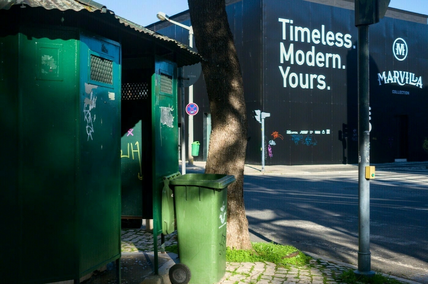 Green public restrooms and a trash bin are situated on a cobblestone sidewalk, with a large advertisement for Marvila Collection on a building in the background.