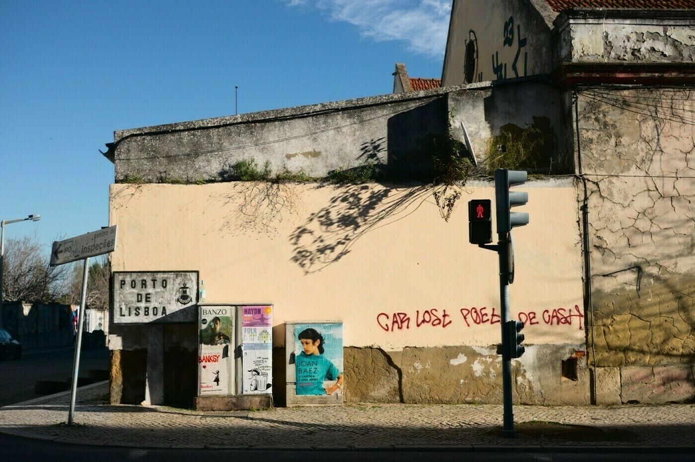 A weathered urban scene features a cracked wall with graffiti and various posters, set against a clear sky and adjacent to a traffic light.