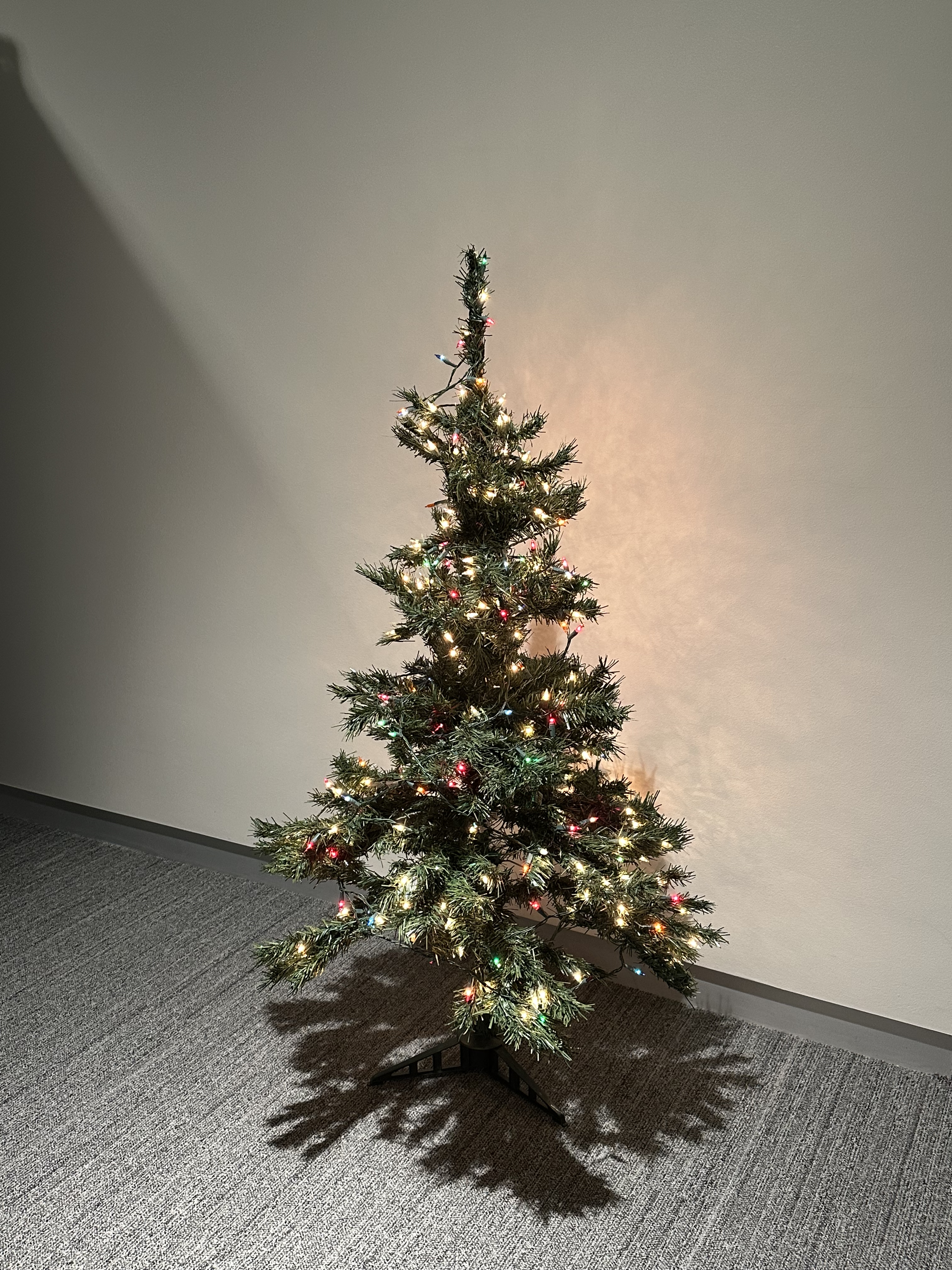 Green, small fake tree lit with white and multicolor lights glows against a white wall