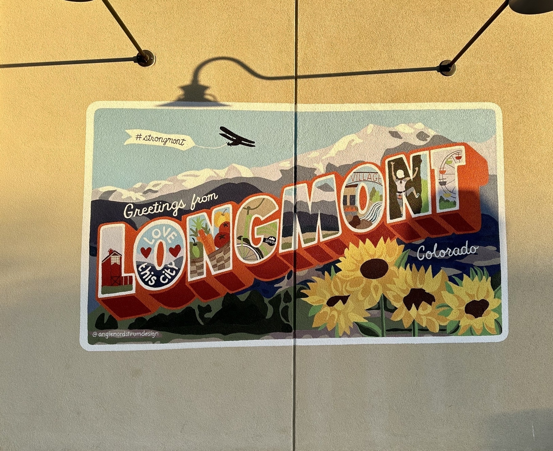 Mural art spelling the city name of "Longmont" Colorado. In each letter is a mini art representing the city. L contains a red barn, O contains "Love this city", N contains a farm basket with veggies, G contains a green bike, M contains mountains, O contains a city center, N contains a person climbing rocks, T contains a ferris wheel