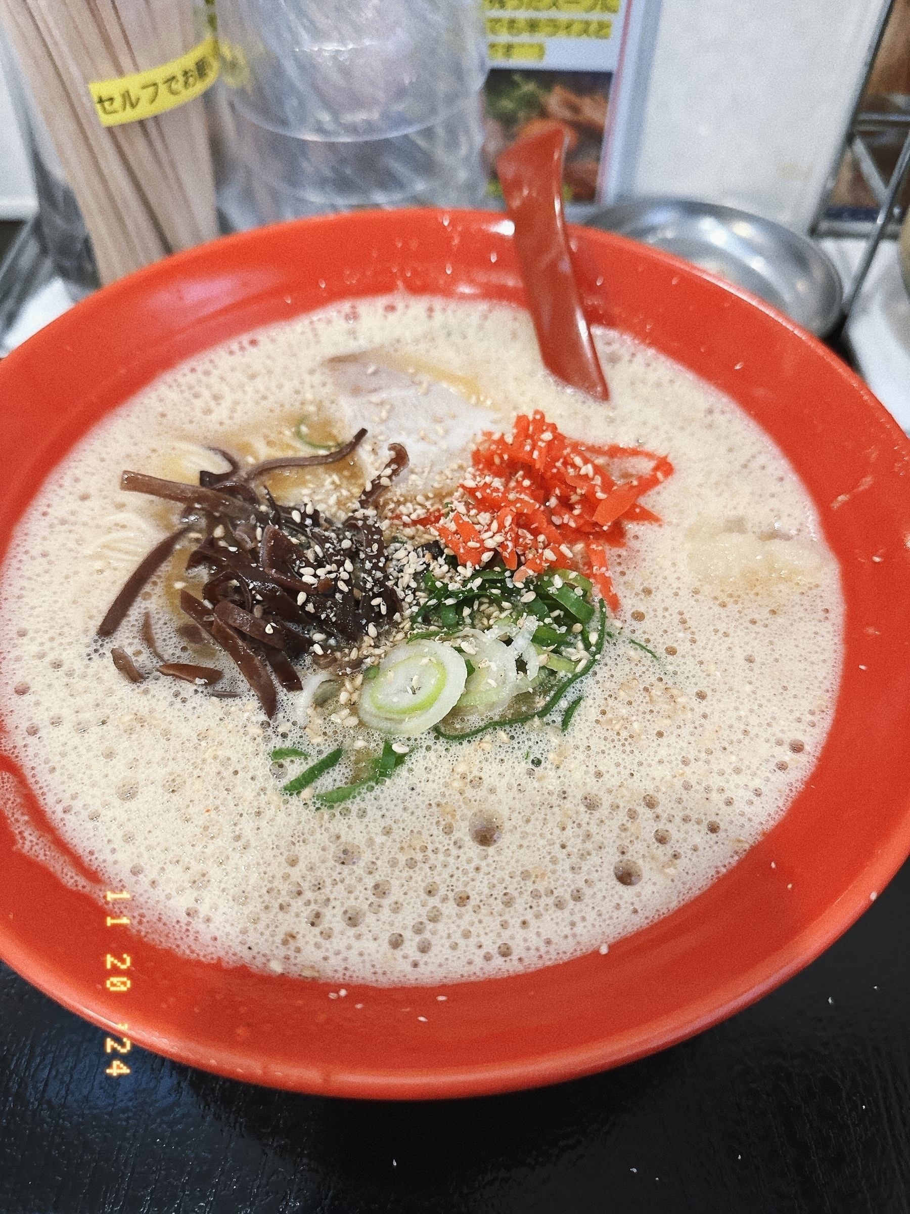 Auto-generated description: A bowl of ramen with various toppings, including sliced green onions, red pickled ginger, and sesame seeds, served in a red bowl.