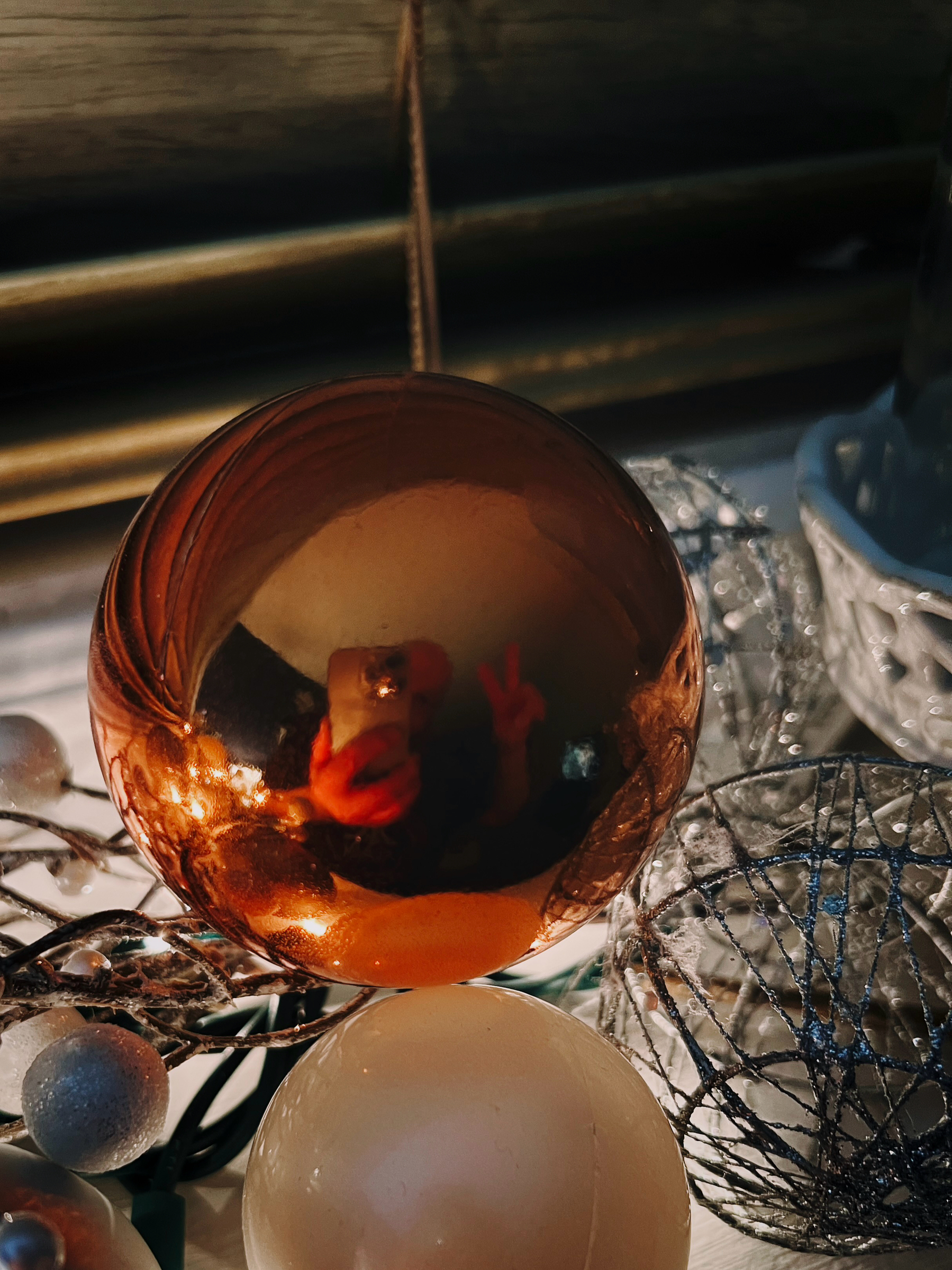 Selfie of my reflection in a spherical orange Christmas ornament, surrounded by white lights, garland, and other smaller ornaments on the window ledge. In my reflection I’m peeking out from behind the phone and giving the peace sign.
