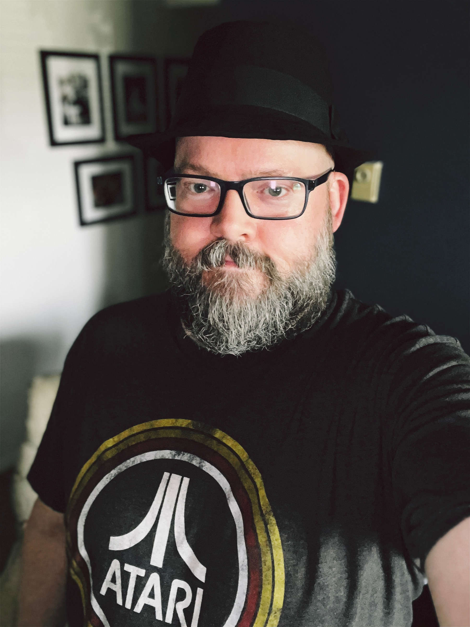 Portrait of me wearing a black Trilby hat, and charcoal-coloured tee with the Atari logo surrounded by concentric circles of white, red, dark yellow, and light yellow.
