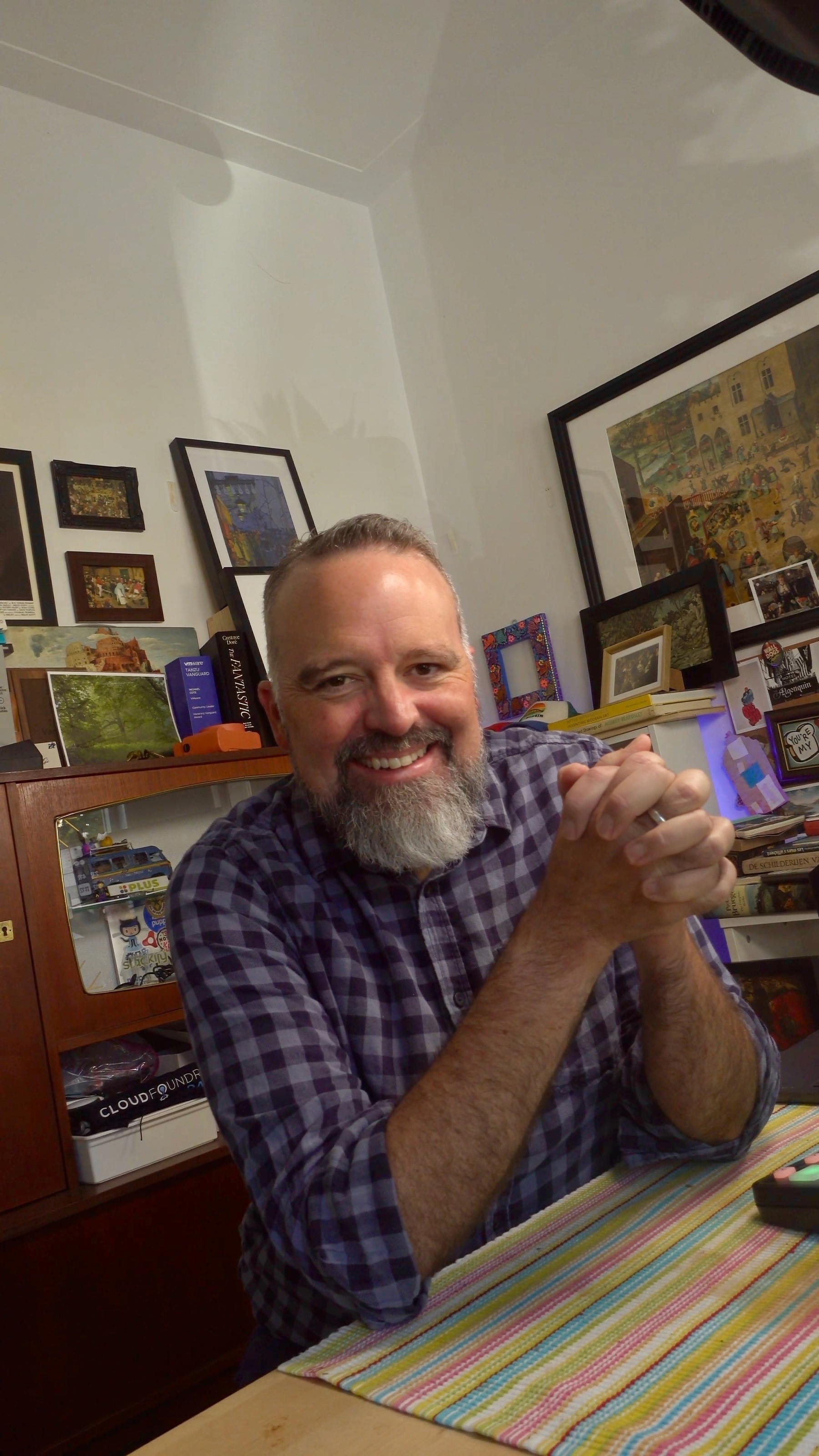 Auto-generated description: A person with a beard is smiling and sitting at a table, surrounded by pictures and books in a cozy room.
