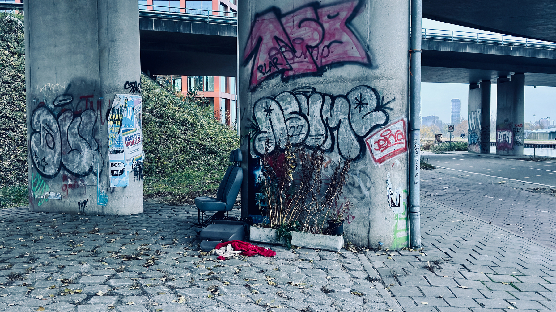 Auto-generated description: Under a highway overpass, graffiti-covered concrete pillars stand alongside scattered chairs and litter, creating an urban, neglected scene.