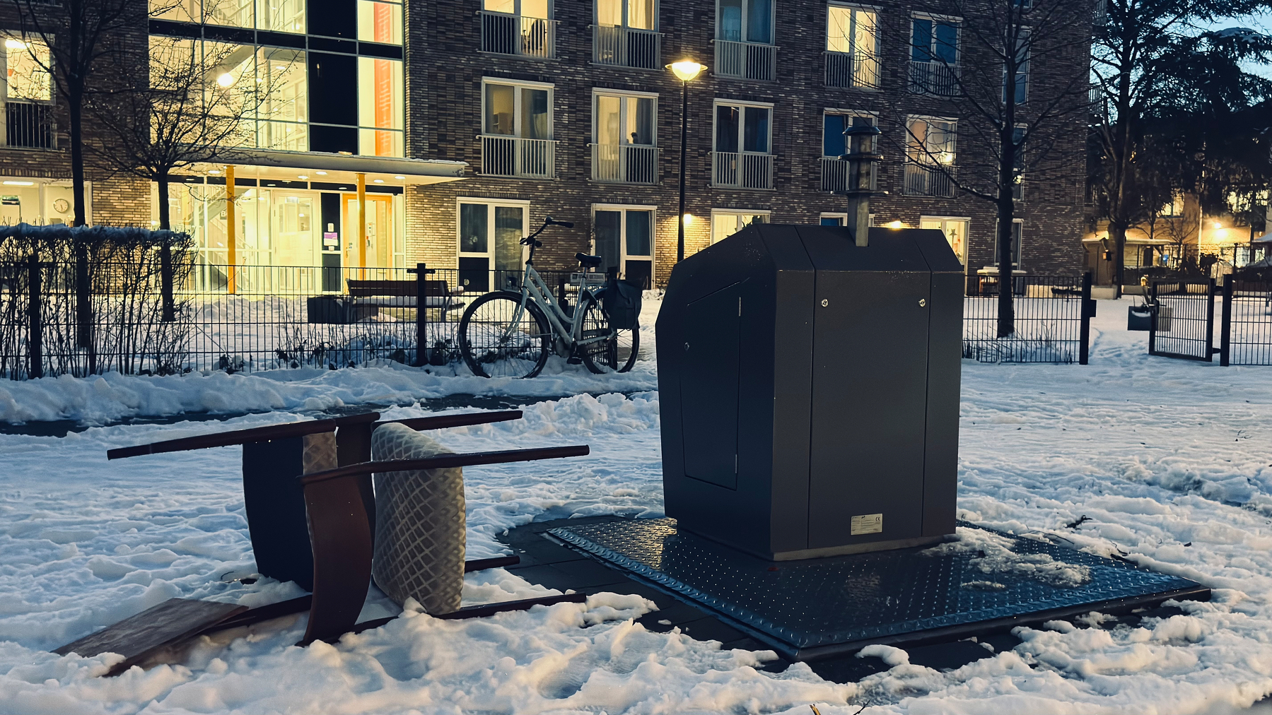 Auto-generated description: A snow-covered outdoor area features an overturned table, a chair, a bike, and a large metal container near an apartment building at dusk.