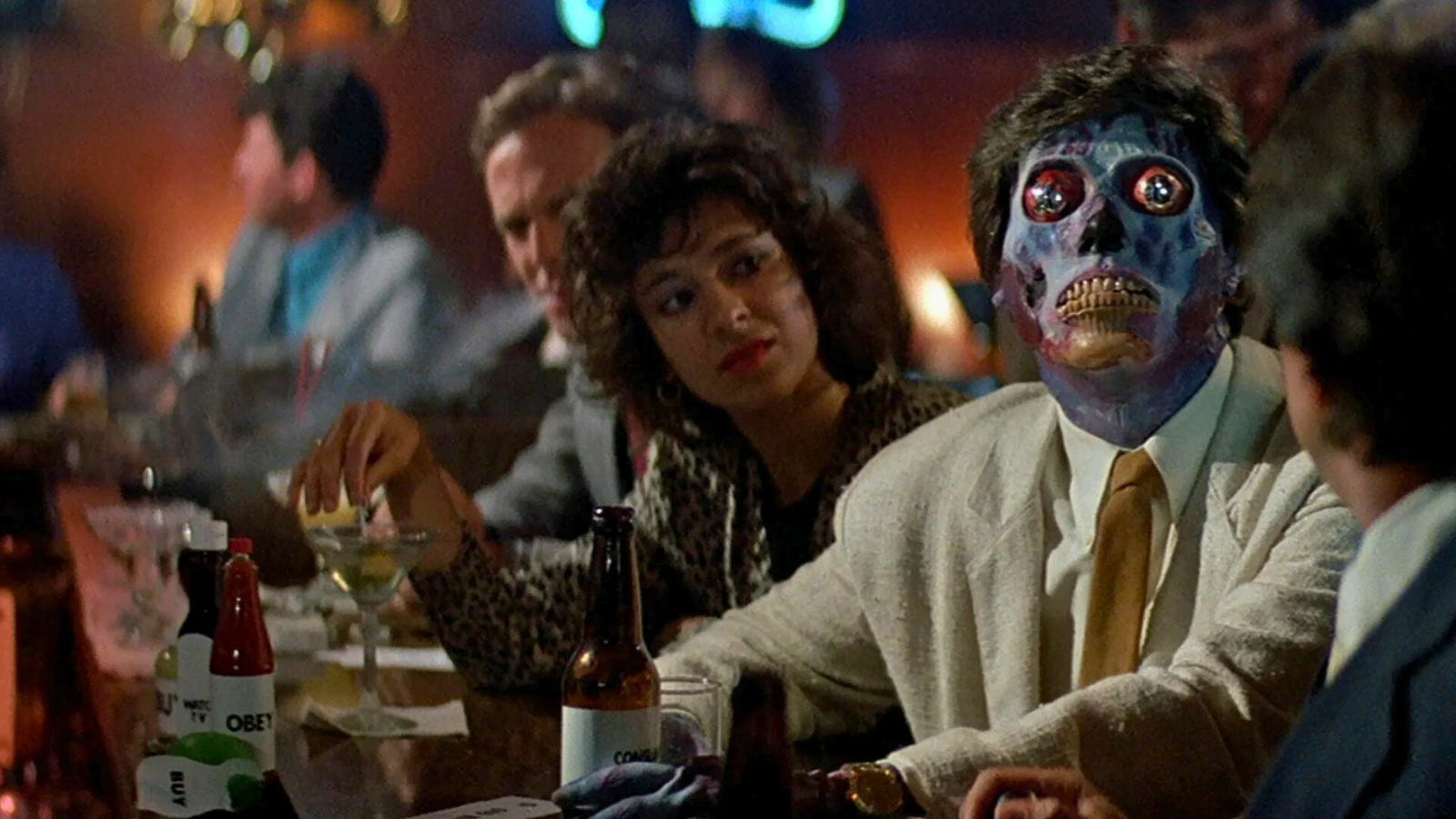 A group of people
  is sitting at a bar, including a person with a half-exposed skeletal face. Still from They Live.