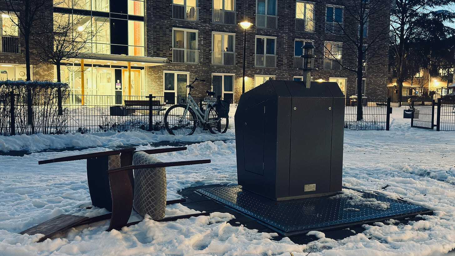 Auto-generated description: A snow-covered outdoor area features an overturned table, a chair, a bike, and a large metal container near an apartment building at dusk.