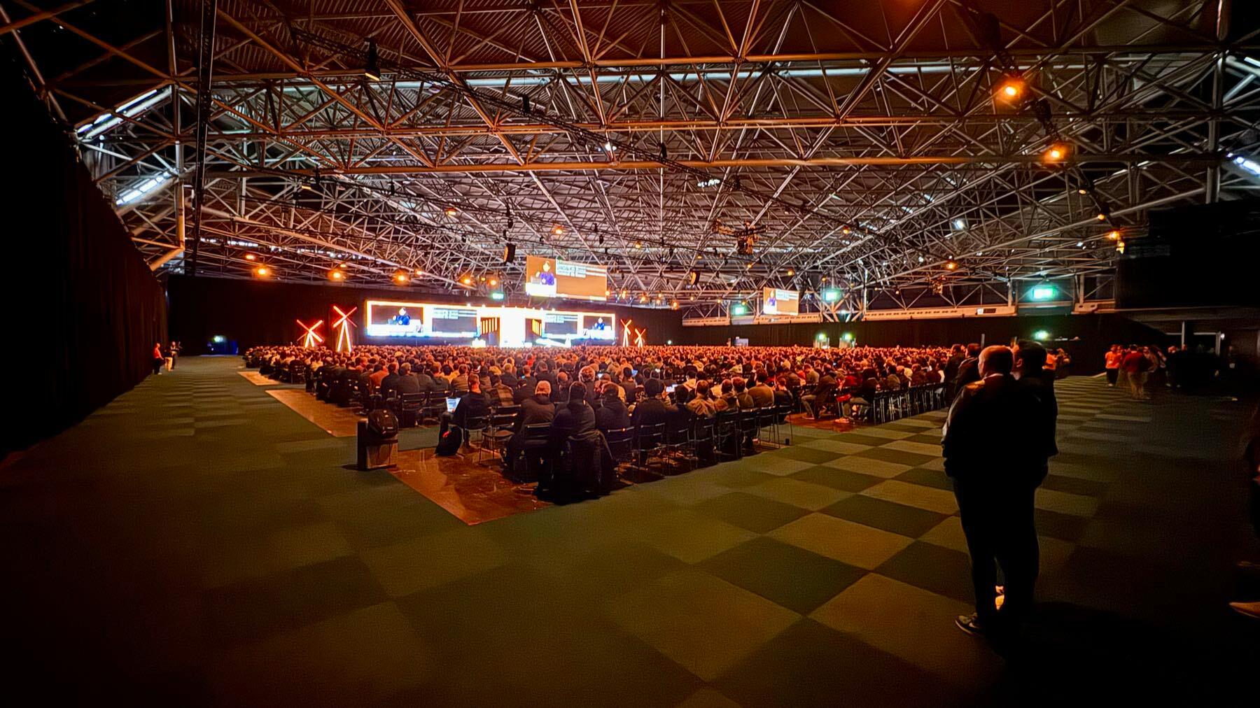 Auto-generated description: A large audience is seated inside a spacious, industrial-style venue with a stage and overhead lighting.
