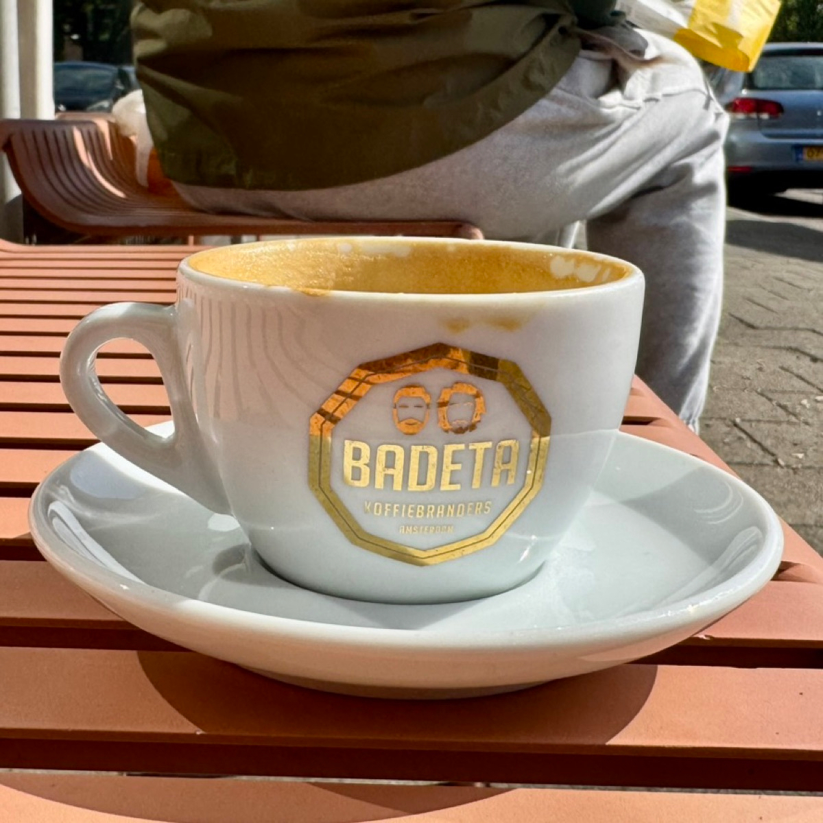 An empty cappuccino cup and saucer on an orange slatted cafe table, the cup printed with a gold Badeta Koffiebranders Amsterdam logo, milk foam dried inside.