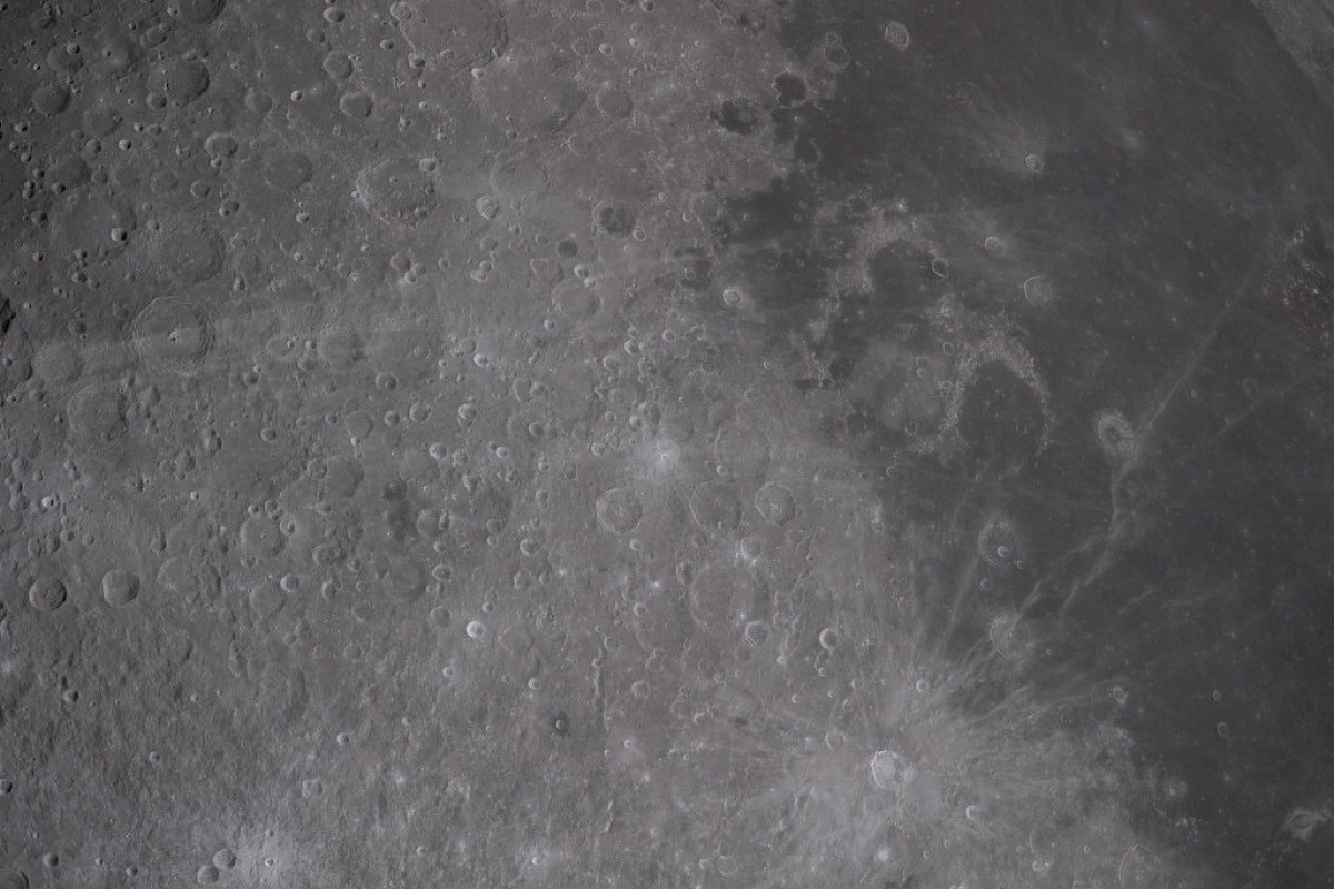Dense black-and-white view of the Moon's cratered surface, overlapping impact craters of many sizes with a prominent ray crater at center-right, bright ejecta streaks radiating outward across the regolith.