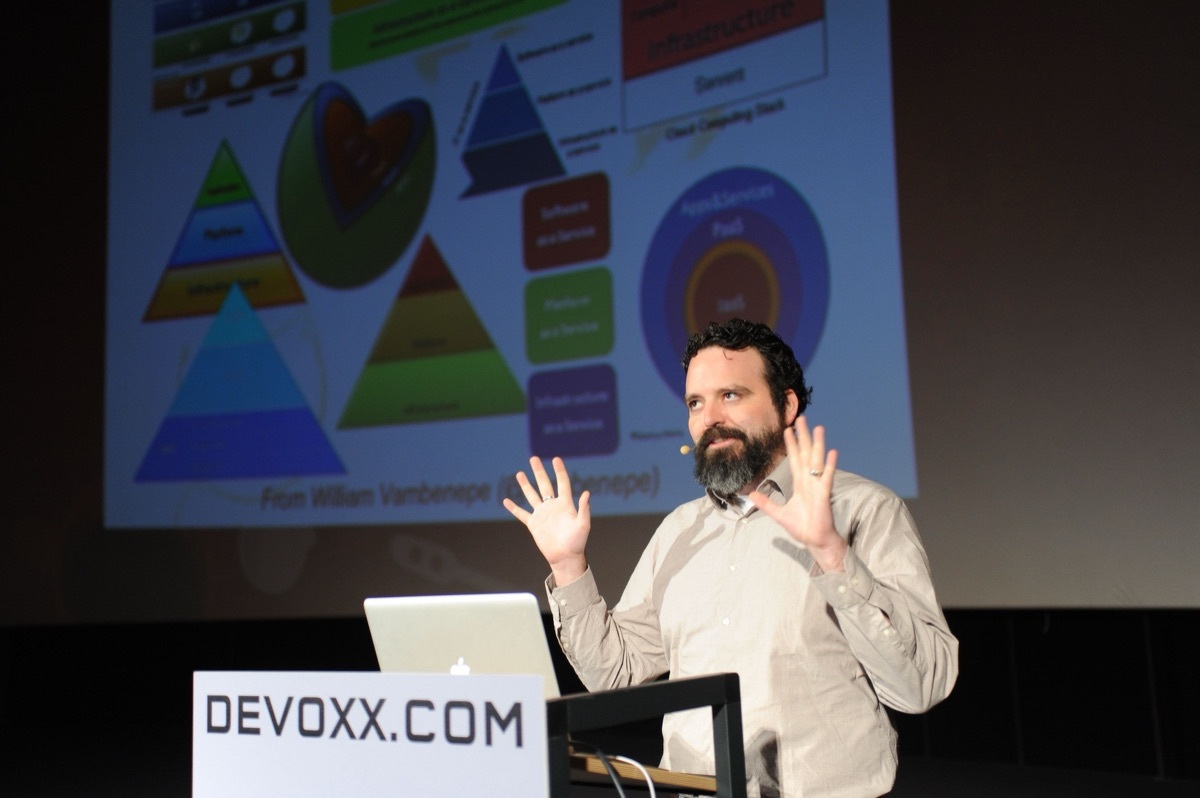 Coté on stage at Devoxx, hands raised mid-gesture, in front of a busy slide of cloud-computing stack pyramids and PaaS/IaaS diagrams.