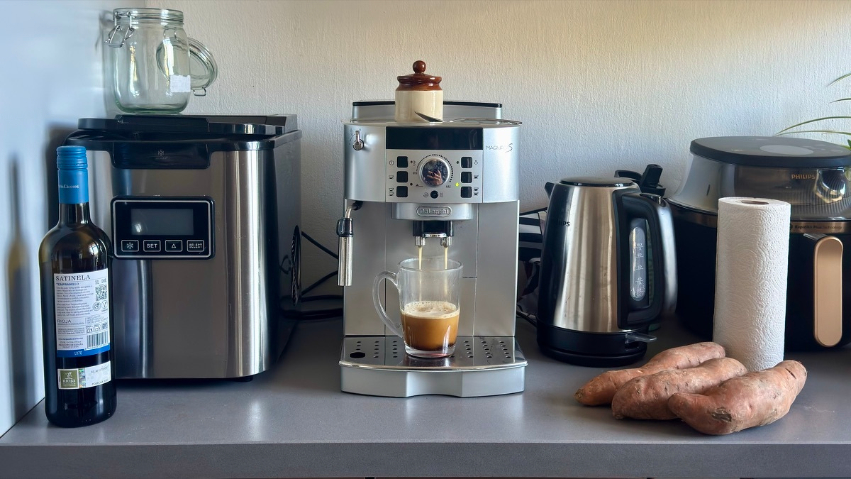 Kitchen countertop still life: a silver De'Longhi espresso machine pulls a fresh shot into a glass mug, flanked at left by a glass storage jar and a bottle of red wine, and at right by a stainless-steel electric kettle, two sweet potatoes, a roll of paper towels, and an air fryer.